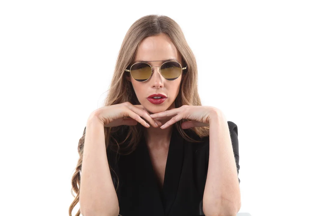 A woman wearing sunglasses and a black outfit posing with her hands under her chin against a white background.